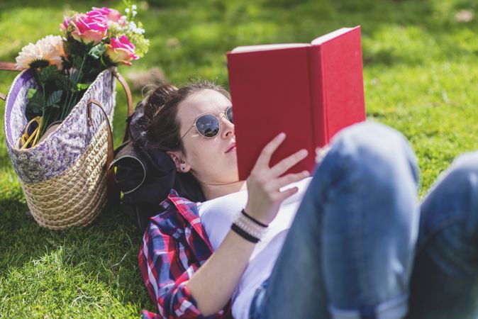 Side view of a hipster happy woman lying on grass in sunny day at park while reading a red book