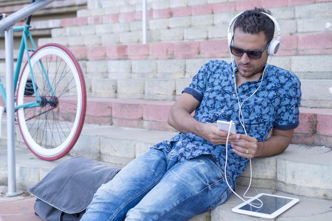 Male cyclist in jeans and blue shirt sitting outside and listening to music on smartphone