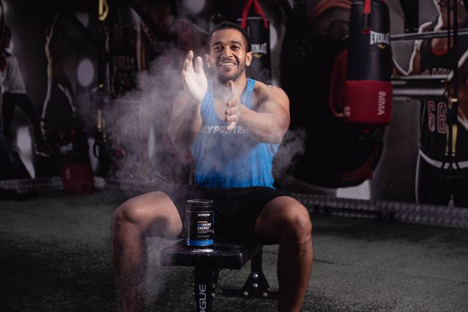 Smiling man spreading powder from protein container sitting on a stool in gym