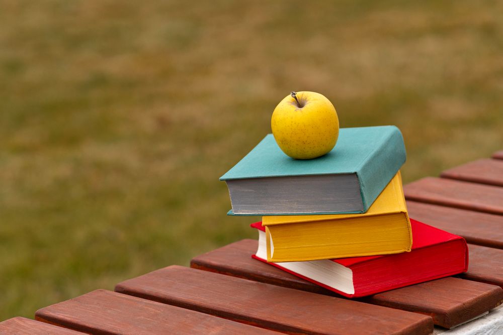 Tilted stack of books with yellow apple placed outdoors.