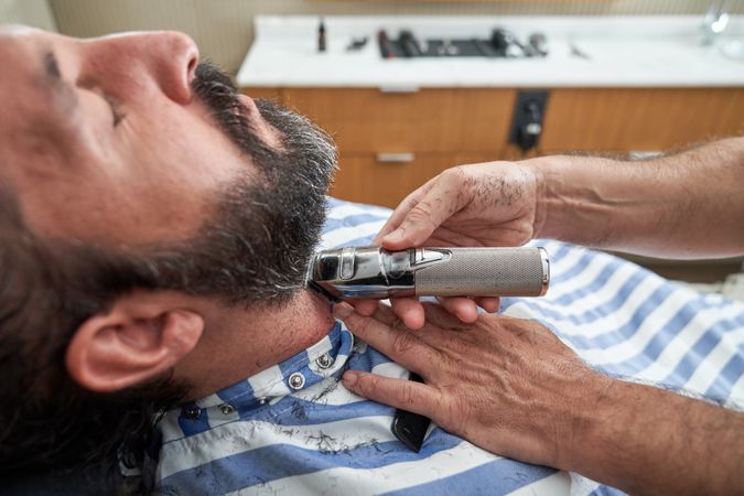 Professional hairdresser trimming beard of male hipster