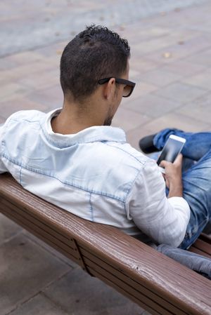 Back of male sitting on bench looking down at phone