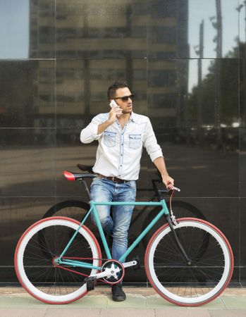 Male standing with his bike next to reflective wall having conversation on smartphone