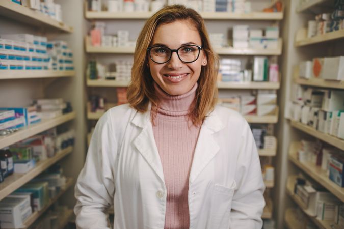 Confident female pharmacist in lab coat standing in pharmacy and looking at camera smiling