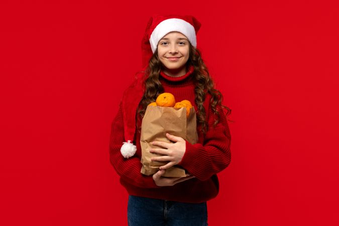 Girl in Santa hat holds paper bag filled with oranges.
