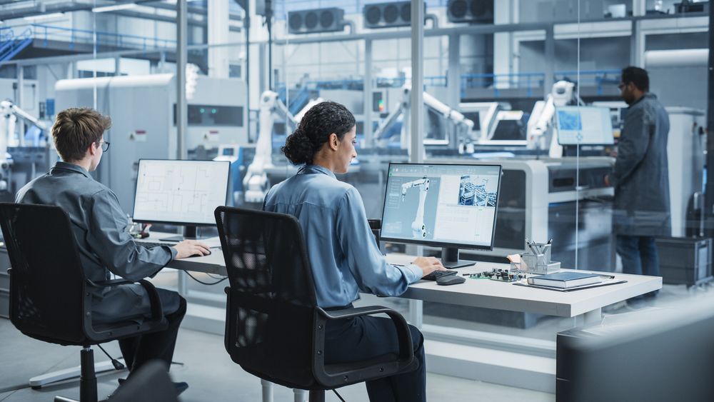 White Male Programmer And Hispanic Female Assembly Line Operator Using Desktop Computers At Modern Factory With Robotic Arms.