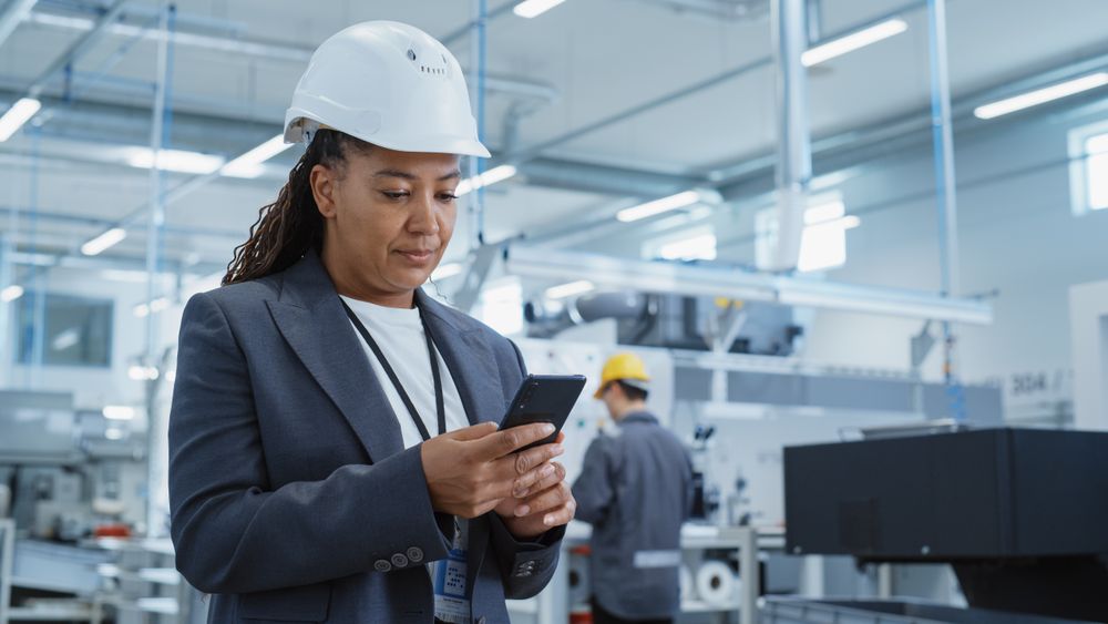 Female Engineer in Hard Hat Standing and Using a Smartphone at Factory.