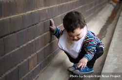 Toddler wearing onesie crouching beside gray wall outdoor 4A9XR4