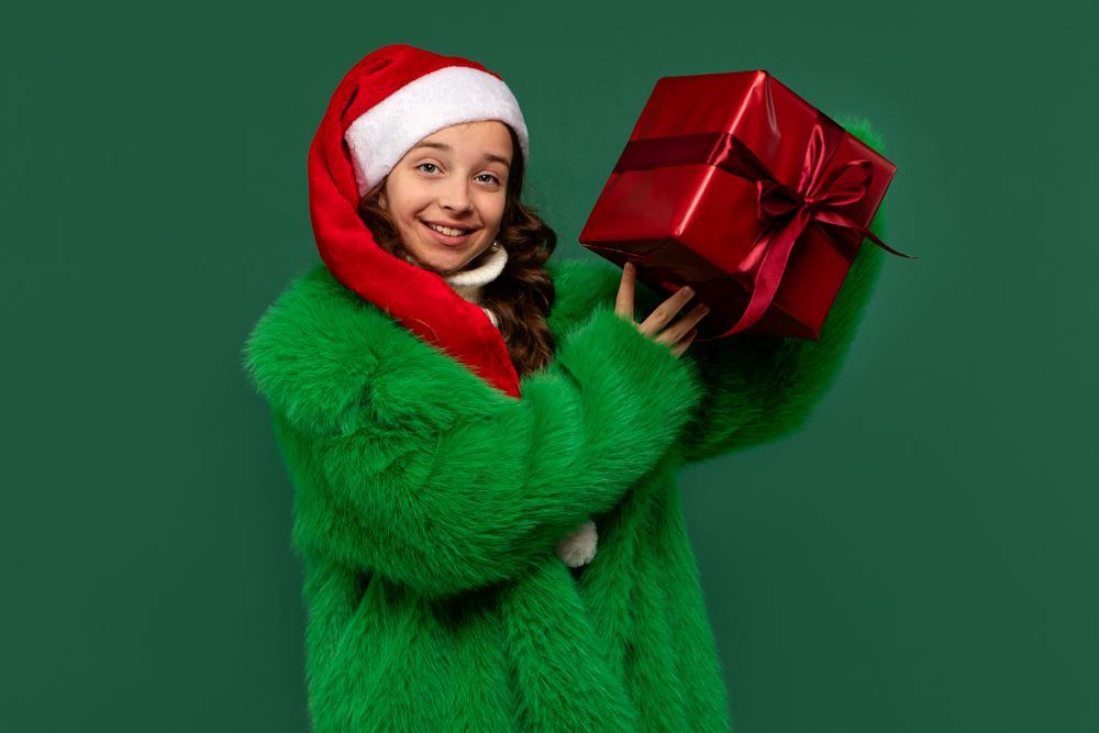 Girl in Santa hat and green fur coat lifting present.