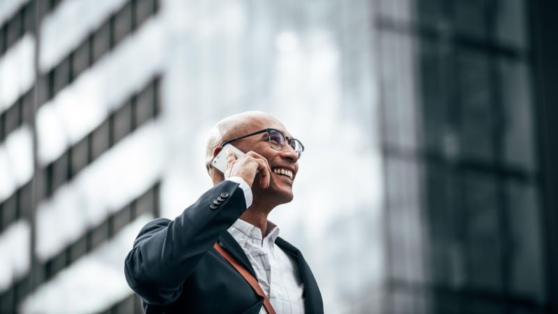 Smiling businessman talking over cell phone while commuting to office with a glass facade building