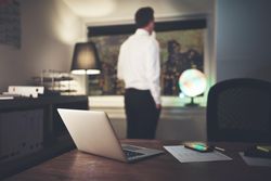 Shot of laptop and cell phone on desk with pensive man in shirt and tie in the background