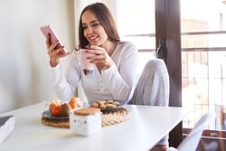 Smiling woman with smartphone eating at table at home