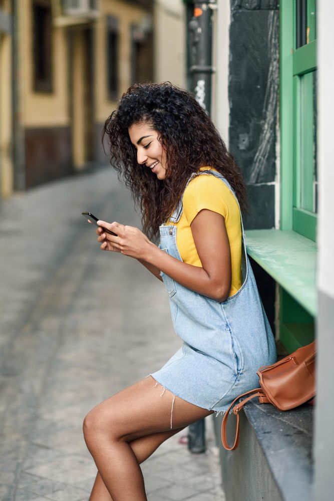 Arab woman perched on green window sill wearing denim dress checking phone