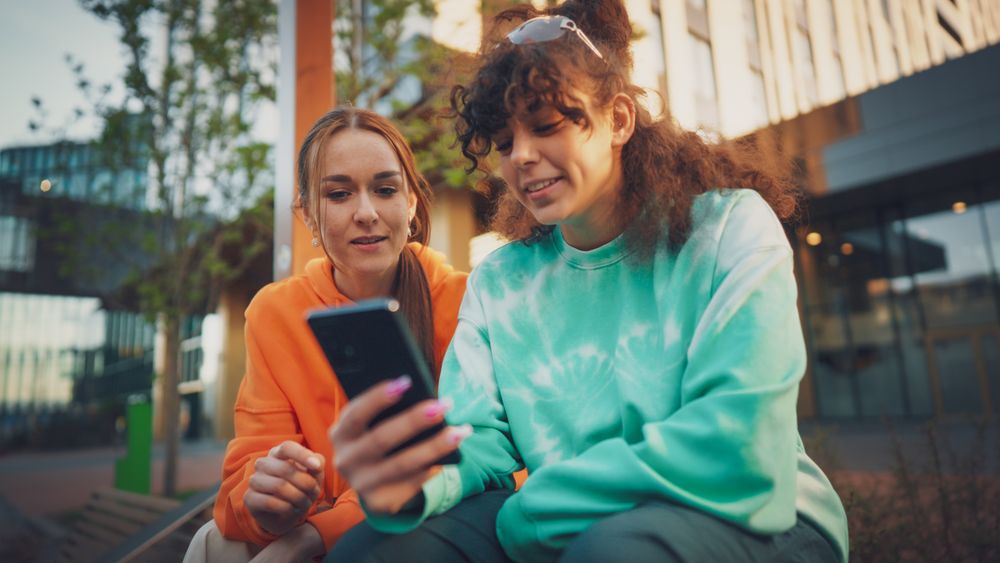 Two Young Women, One With Curly Hair In A Vibrant Tie-dye Sweatshirt, Share A Moment Of Joy