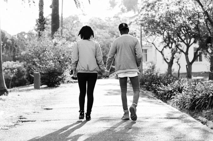 Back view of man and woman holding hands and walking on sidewalk in grayscale