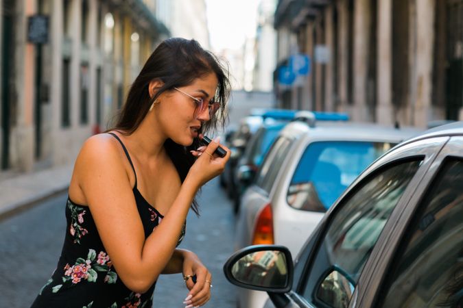 Confident woman in sunglasses applying lipstick while looking at car window on street