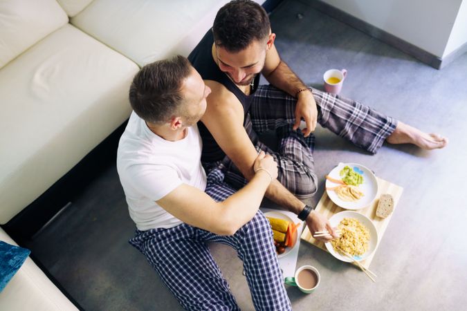 Looking down at male couple eating meal together at home sitting on living room floor