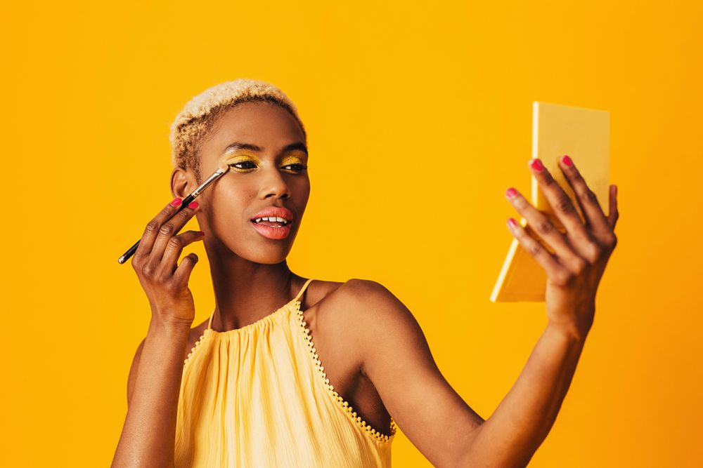 Black woman with short blonde hair holding a pocket mirror applying gold eyeshadow