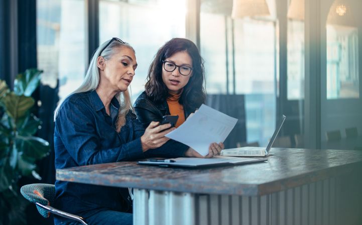 Woman executive showing her phone and papers to younger colleague at office