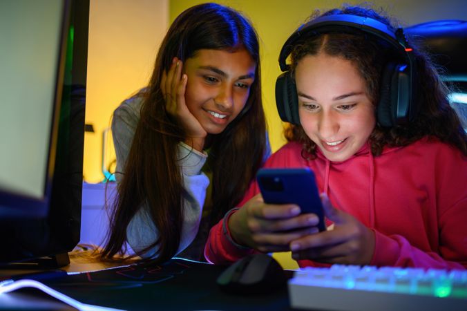 Happy multiracial teenage girlfriends watching smartphone in room with gaming PC