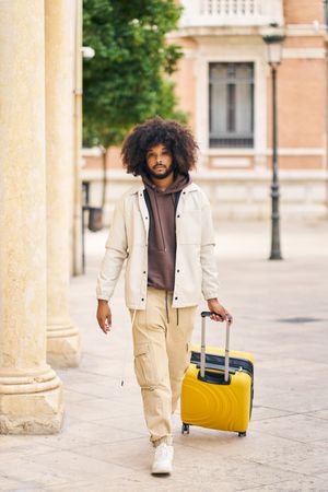 Portrait of a young man with afro hair walking down the street with yellow suitcase