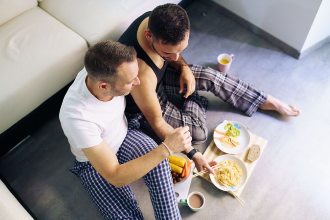 Looking down at male couple enjoy pasta and vegetables at home sitting on floor