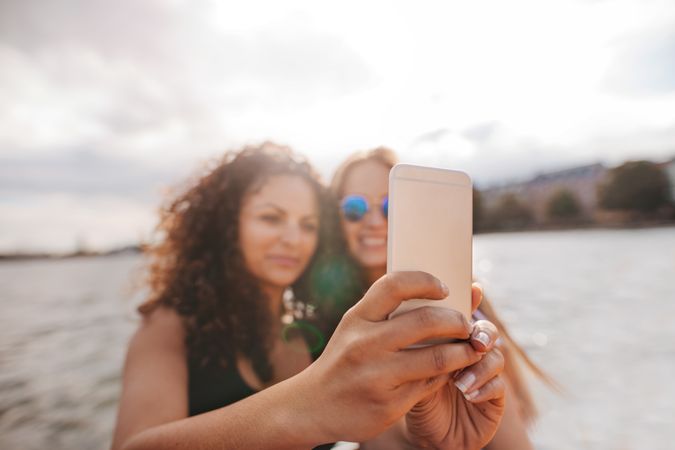 Two female friends taking selfie on the water with focus on smart phone