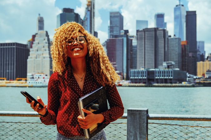 Smiling professional Black female holding her phone and notebooks by Hudson River