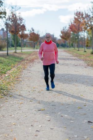 Grey haired man in red shirt jogging on in tree lined park