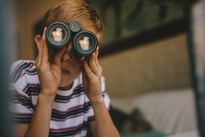 Small boy looking outside window using binoculars