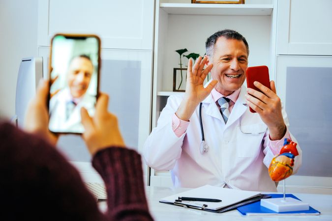 Smiling physician waving on video call with patient from his office