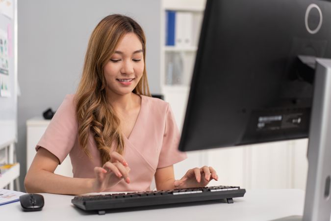 Happy Asian woman sitting in office and concentrating on typing on keyboard