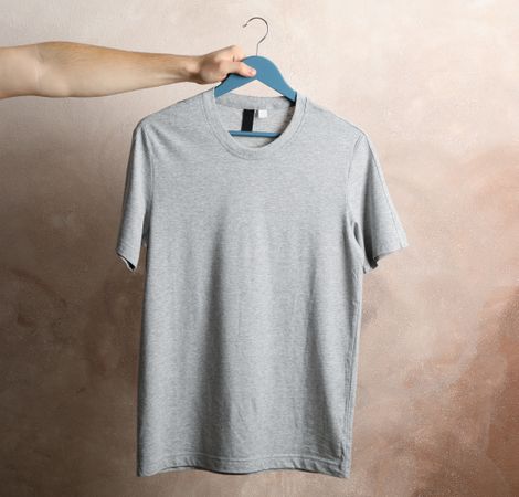Man holds a hanger with blank gray t-shirt on brown background, space for text