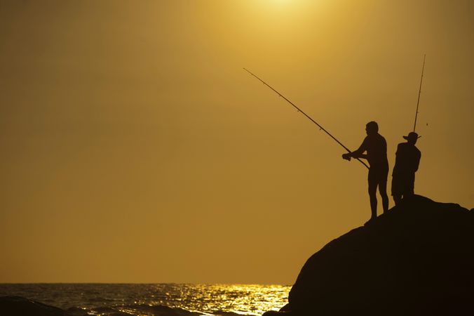 Silhouette of men fishing by shoreline at sunset