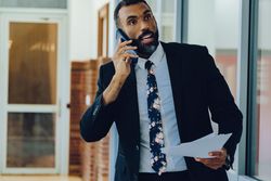 Businessman in suit and tie with papers walking in office while speaking on smart phone