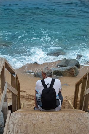 Back view of middle aged man with backpack sitting on wooden stairs by the sea
