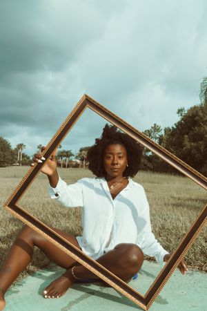 Black woman sitting on brown grass holding brown wooden frame