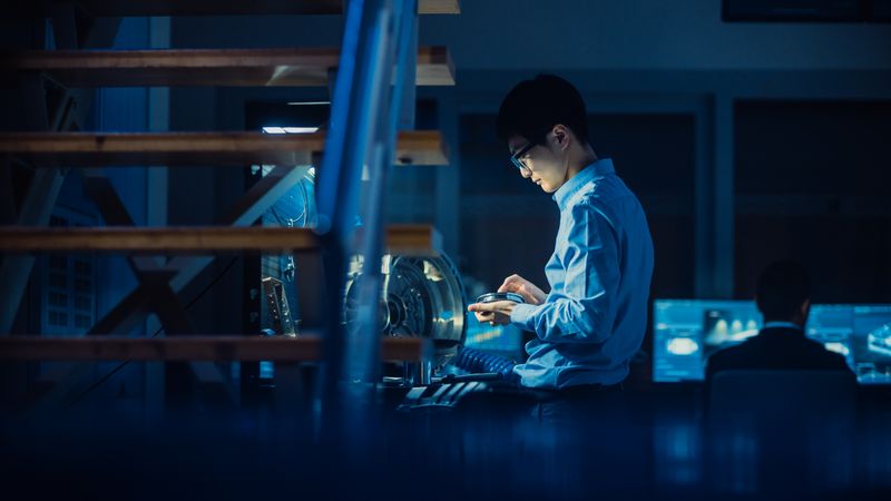 Professional Automotive Engineer in Glasses is Working on Transmission Gears in a High Tech Innovative Laboratory with a Computer Screens.