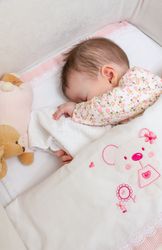 Baby girl covering her face, asleep in a cot with pacifier and toy