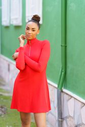 Smiling female in red zipped up dress standing outside green building touching her face