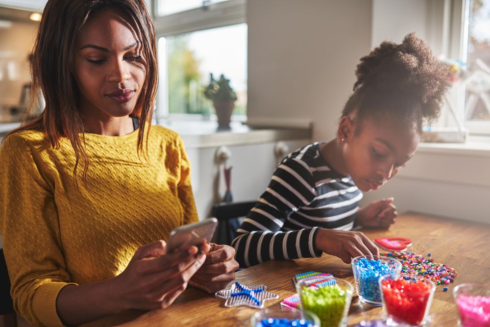 Black mother checks phone with daughter doing arts and crafts at home