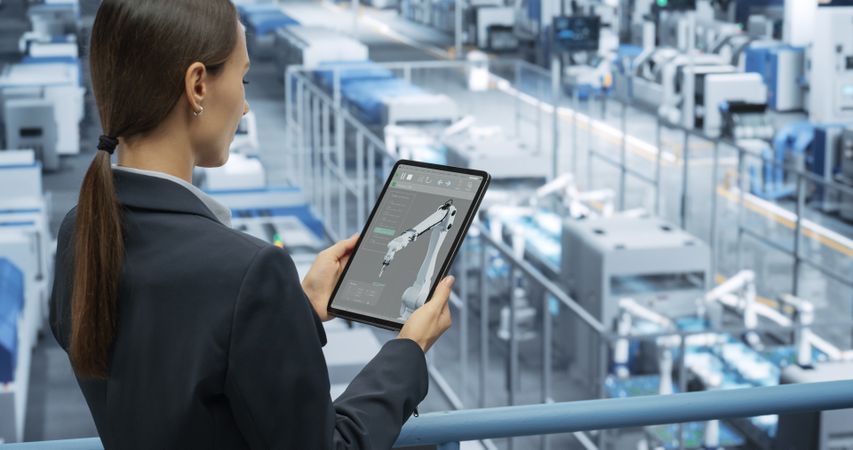 Portrait of a Happy Engineer Using Laptop Computer and Looking Around a Factory Facility with Automated Robotic Arms Producing Modern Electronic Components for Different Industries