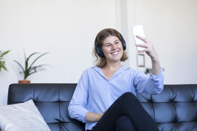 Woman  in headphones sitting on a sofa taking selfie on her phone