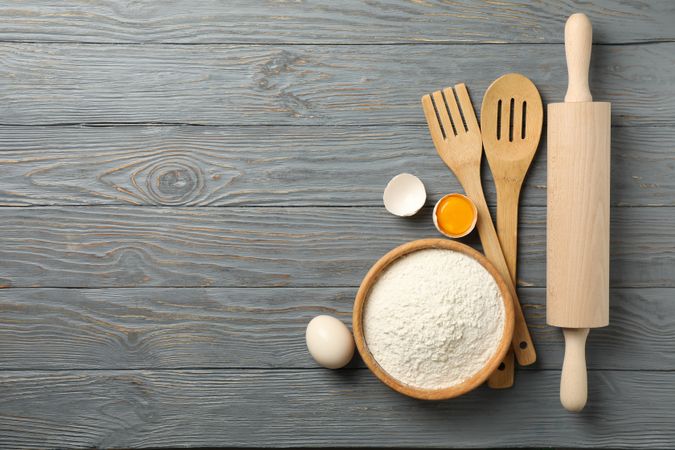 Kitchen cutlery, flour and eggs on gray wooden background