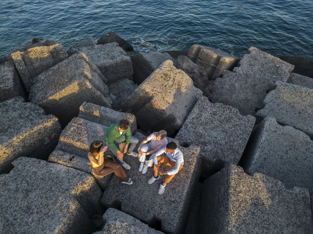 Aerial view of diverse friends sitting on rocks by the sea at sunset