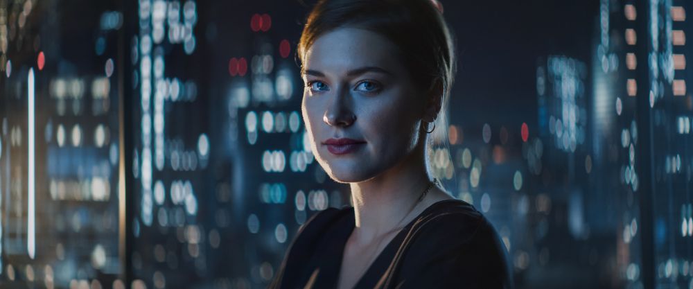 Close Up Portrait of Businesswoman Posing Next to Window in Big City Office with Skyscrapers Late At Night.