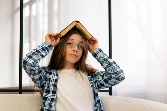 Girl holding book over head sitting on sofa indoors.