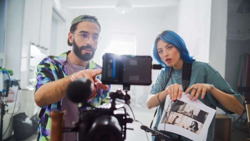 In A Bright Studio, A Young Male And Female With Blue Hair Review Footage On A Digital Camera, Surrounded By Filmmaking Equipment, Embodying Creativity And Collaboration In Movie Production.