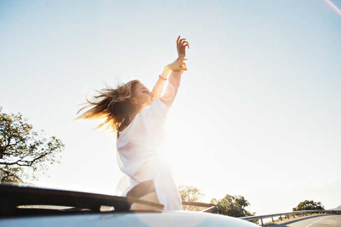 Rearview of woman waving out of sunroof on sunny day with her arms up