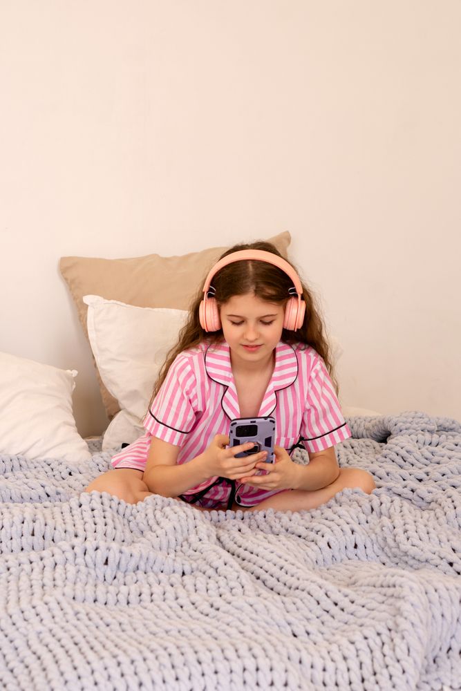 Girl in pink striped pajamas using smartphone on bed.